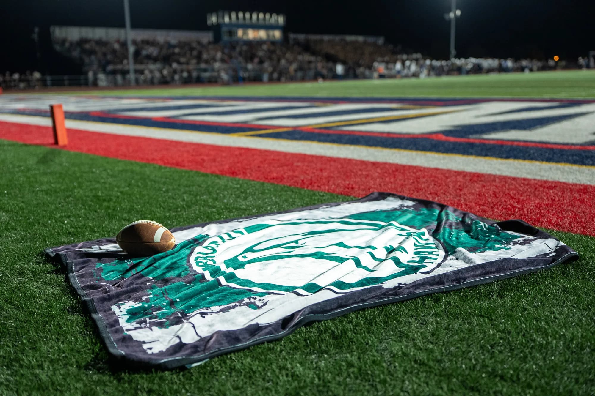 A custom school blanket and a football laid on the endzone turf under the lights, a full night crowd in the stands behind