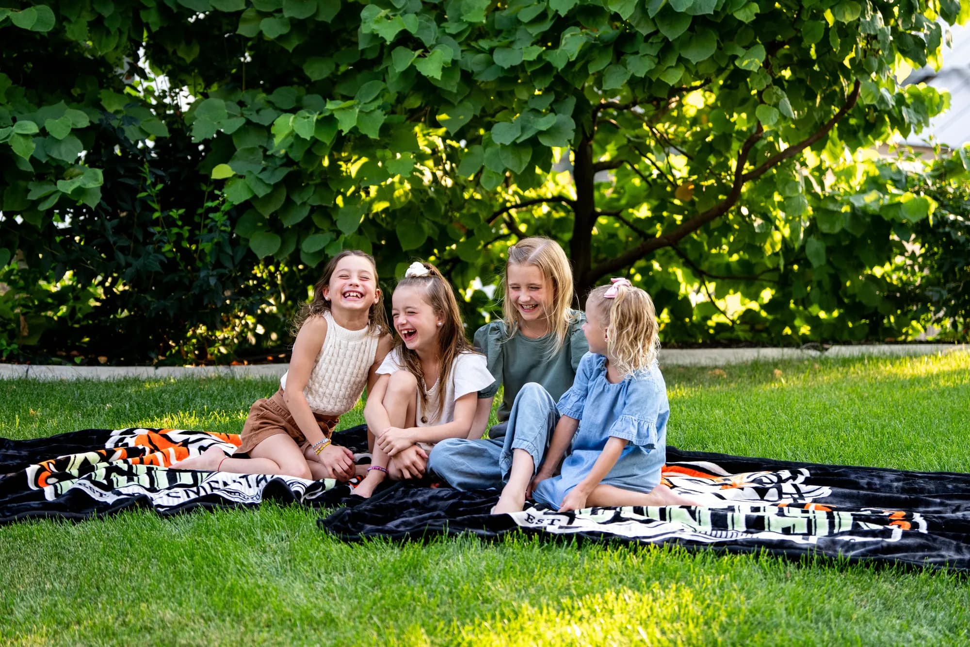 Four kids sitting and laughing together on custom school blankets spread across a sunny lawn