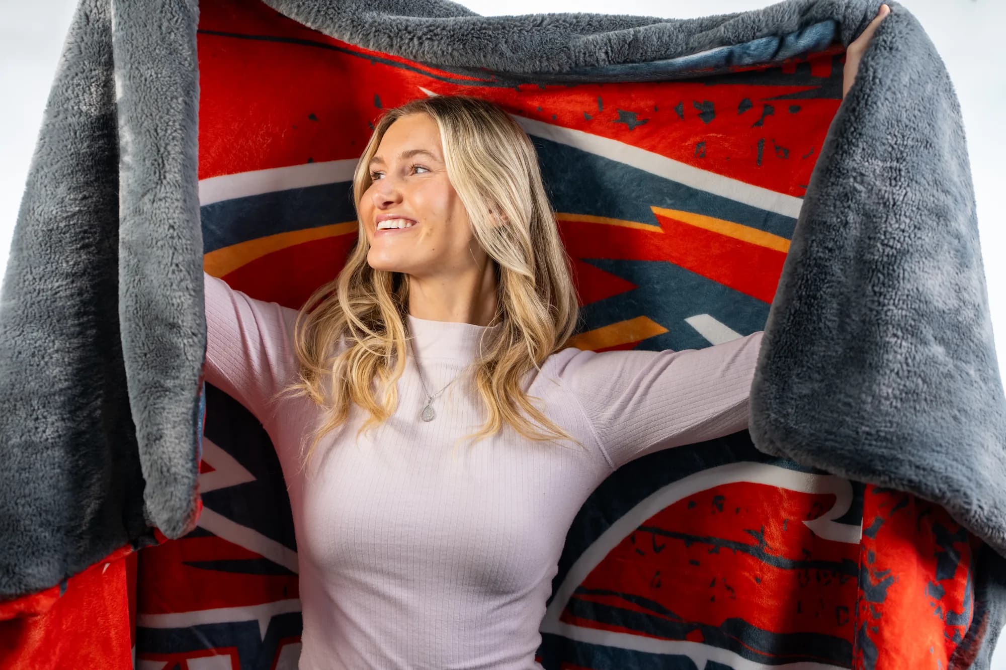 A young woman holds up the Swanky Blanky, plush gray faux fur on the back and a custom red-and-navy NubaySoft 2.0 school print on the front